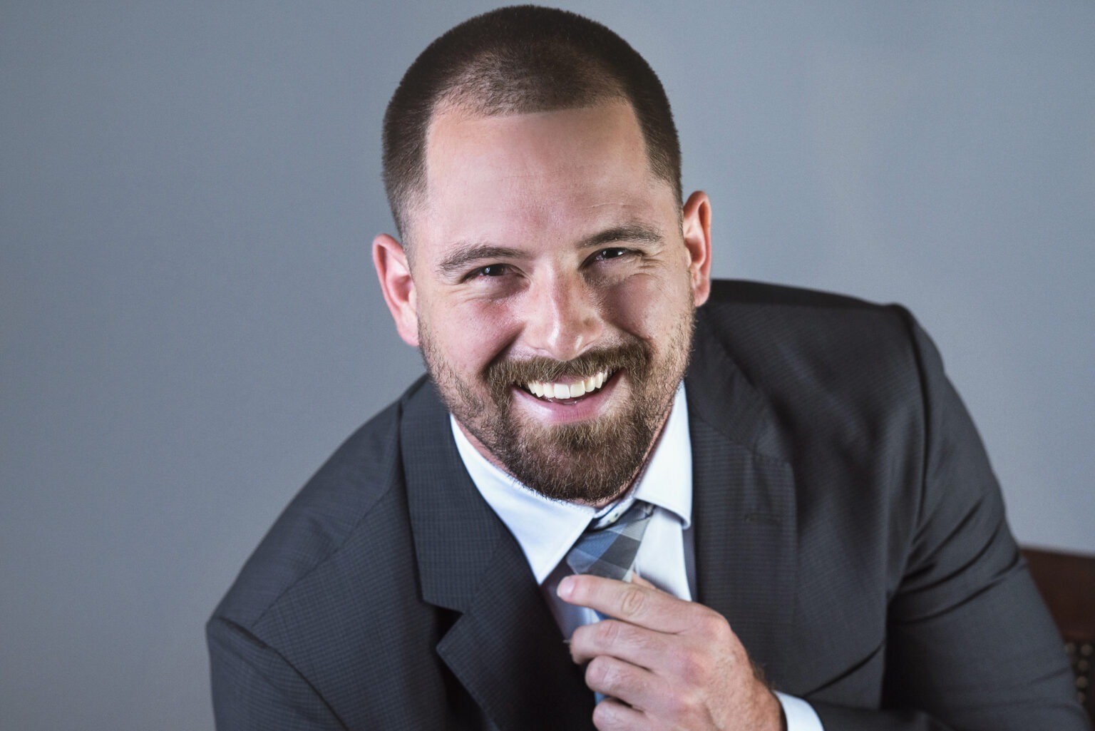Professional studio headshot of a young man in a dark suit adjusting his tie and smiling confidently. Captured by David Scarola Photography in Palm Beach County, Florida.professional headshot photographer Palm Beach, corporate portraits Jupiter Island, studio headshots South Florida, business portrait photographer Miami, LinkedIn headshots Palm Beach County, modern executive portrait Florida, professional branding photography, David Scarola Photography, headshot photography for young professionals, best headshot photographer Palm Beach and Miami.