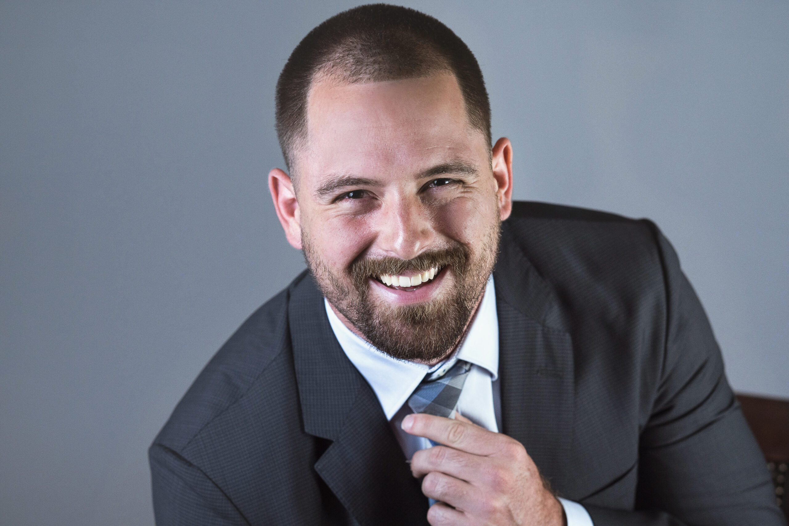 Professional studio headshot of a young man in a dark suit adjusting his tie and smiling confidently. Captured by David Scarola Photography in Palm Beach County, Florida.professional headshot photographer Palm Beach, corporate portraits Jupiter Island, studio headshots South Florida, business portrait photographer Miami, LinkedIn headshots Palm Beach County, modern executive portrait Florida, professional branding photography, David Scarola Photography, headshot photography for young professionals, best headshot photographer Palm Beach and Miami.