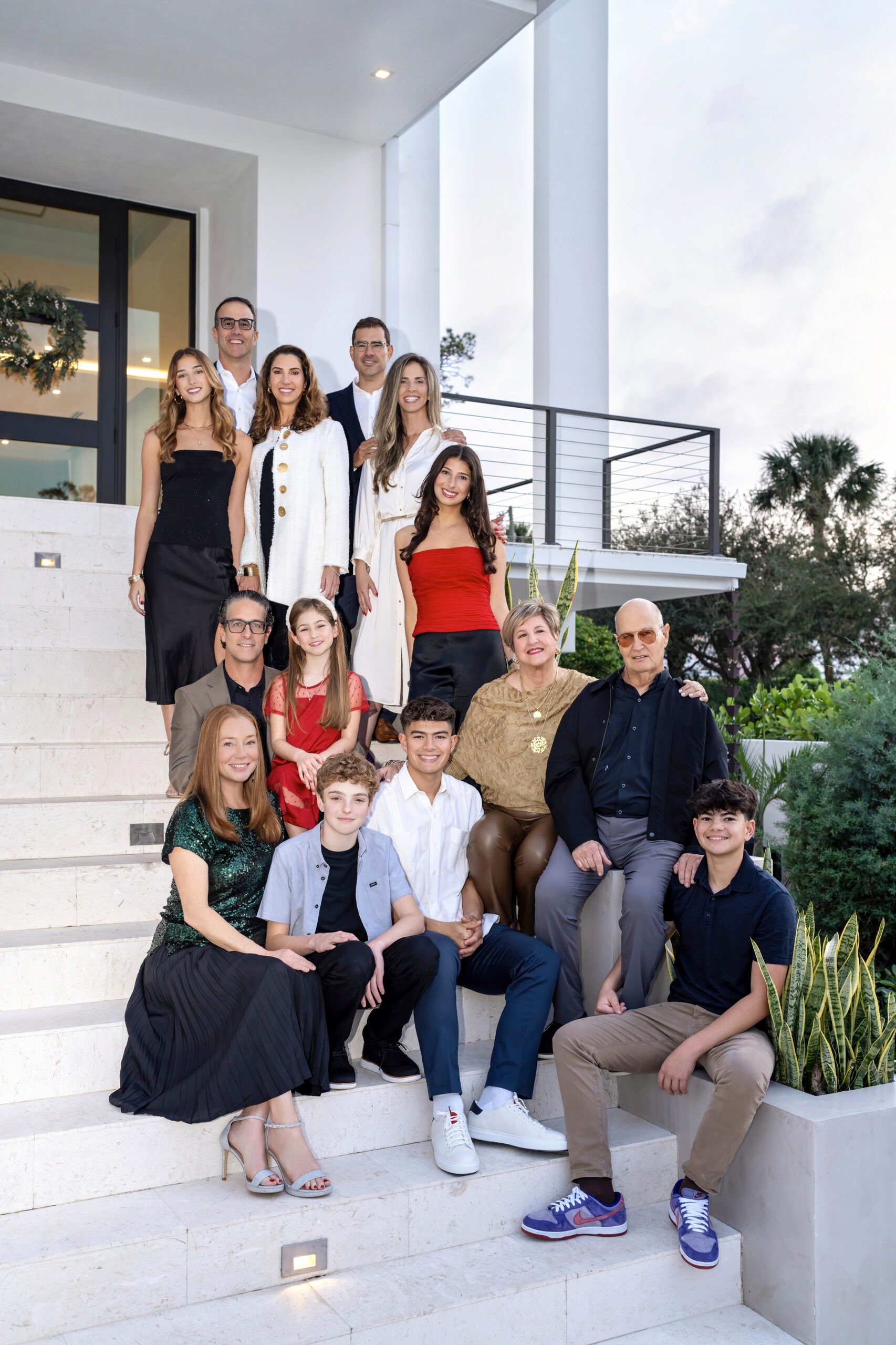 A large extended family poses together on the steps of a modern Palm Beach home, dressed in elegant holiday attire. The group includes multiple generations—grandparents, parents, and children—all smiling warmly during a professional family photography session.family photographer Jupiter Island, best portrait artist USA, Palm Beach family photography, professional family portraits, fine art photographer Florida, family event photographer, holiday portraits Palm Beach, master photographer, David Scarola Photography, autism-friendly photographer, multi-generational family portraits