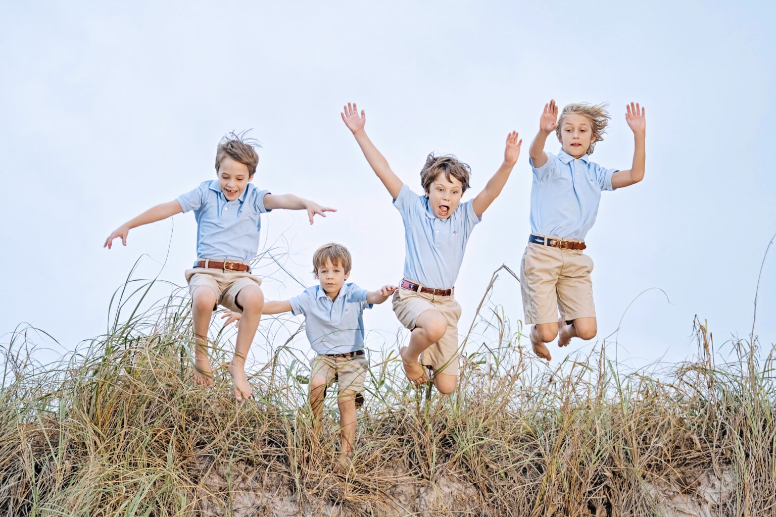 Four brothers in matching blue shirts and khaki shorts jumping off the sand dunes on Palm Beach Island, captured midair with joyful expressions by David Scarola Photography — Palm Beach’s top family photographer.