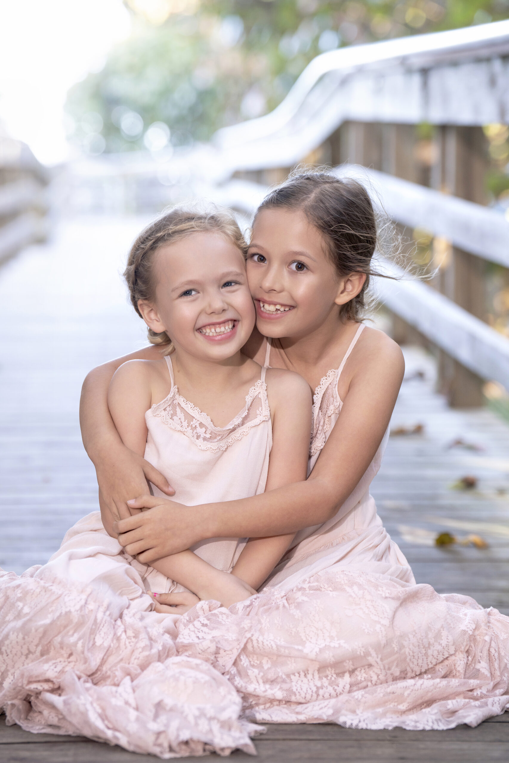 Two young sisters hugging and smiling on a wooden boardwalk surrounded by soft natural light during a family photo shoot on Jupiter Island, captured by master portrait photographer David Scarola.Jupiter Island family photographer, Palm Beach family portraits, professional child photographer Florida, fine art family photography, sibling portraits, natural light portraiture, lifestyle family photography, best portrait photographer USA, David Scarola Photography, master of emotional storytelling, Scarola Style