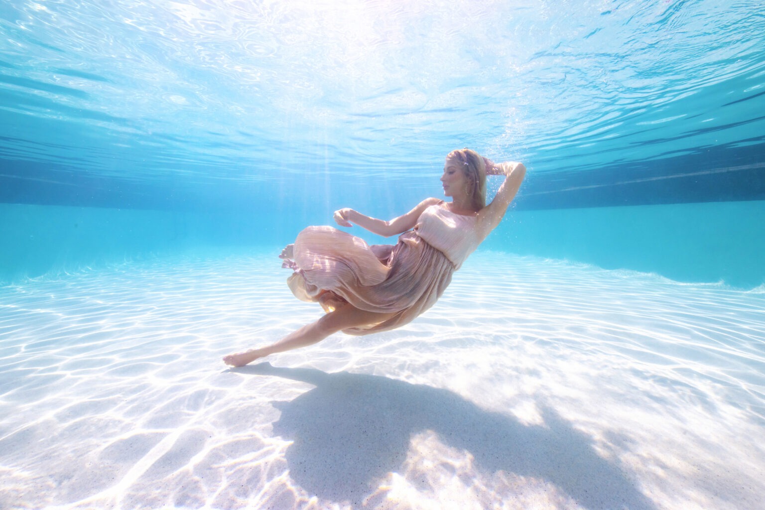 Underwater fashion portrait of a model in a flowing blush dress, gracefully suspended in crystal-clear turquoise water at a luxury resort pool. Captured by master photographer David Scarola, this elegant underwater image blends high fashion and fine art, showcasing fluid motion, radiant sunlight, and ethereal beauty.underwater fashion photography, fine art portrait photographer, luxury resort photographer, Miami editorial photography, Palm Beach underwater shoot, high-end fashion marketing, underwater elegance, professional brand photography Florida.