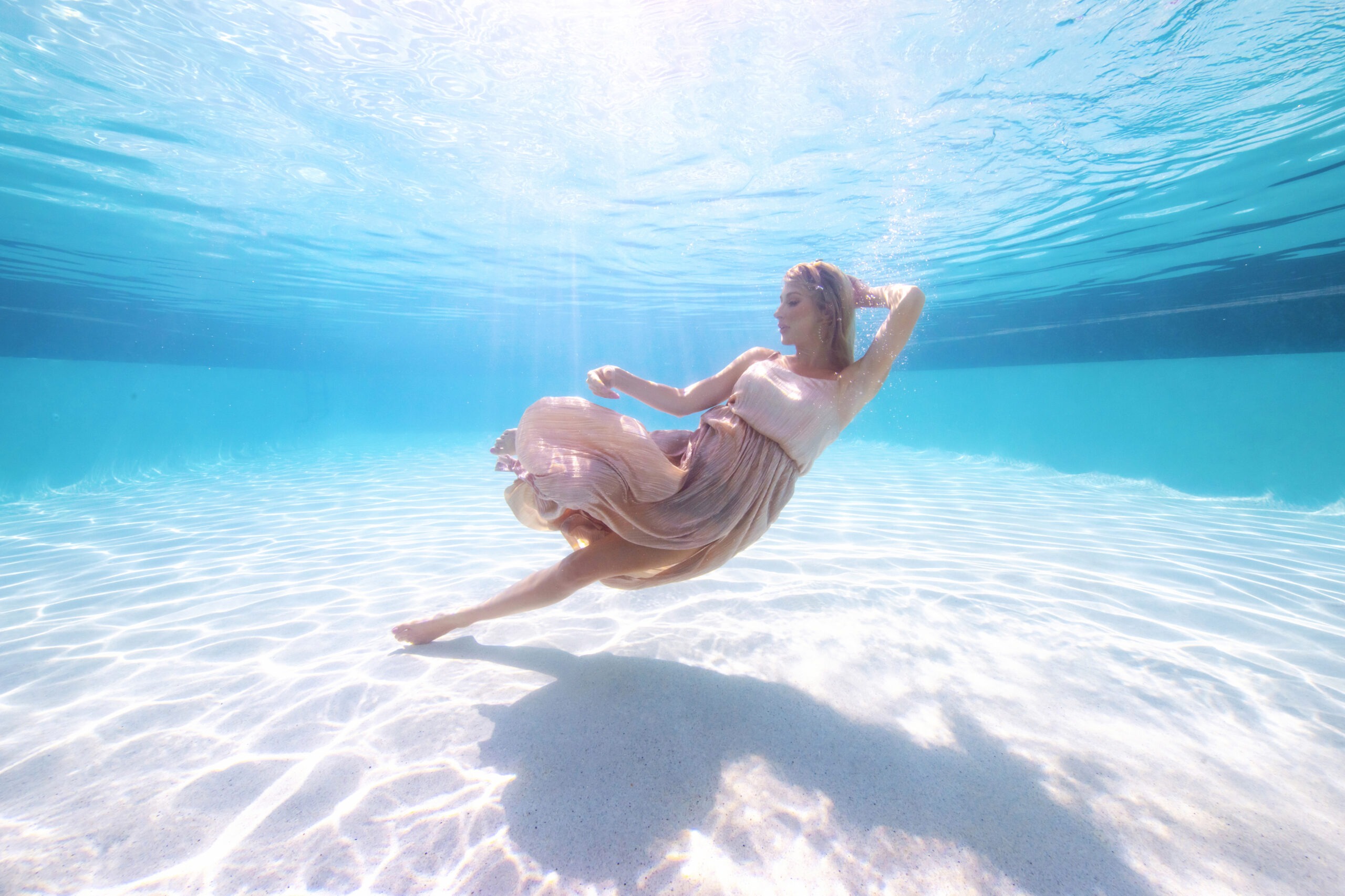 Underwater fashion portrait of a model in a flowing blush dress, gracefully suspended in crystal-clear turquoise water at a luxury resort pool. Captured by master photographer David Scarola, this elegant underwater image blends high fashion and fine art, showcasing fluid motion, radiant sunlight, and ethereal beauty.underwater fashion photography, fine art portrait photographer, luxury resort photographer, Miami editorial photography, Palm Beach underwater shoot, high-end fashion marketing, underwater elegance, professional brand photography Florida.