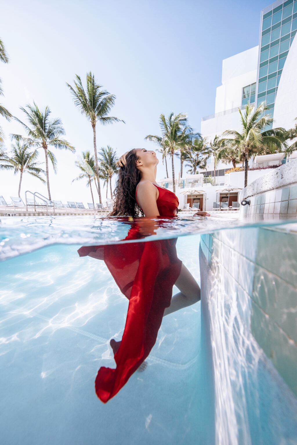 A woman in a flowing red dress rises gracefully from a luxury resort pool under bright blue skies, surrounded by palm trees and modern architecture at The Ritz-Carlton Fort Lauderdale. Captured half above and half below water by David Scarola Photography, blending fine art, fashion, and resort elegance.