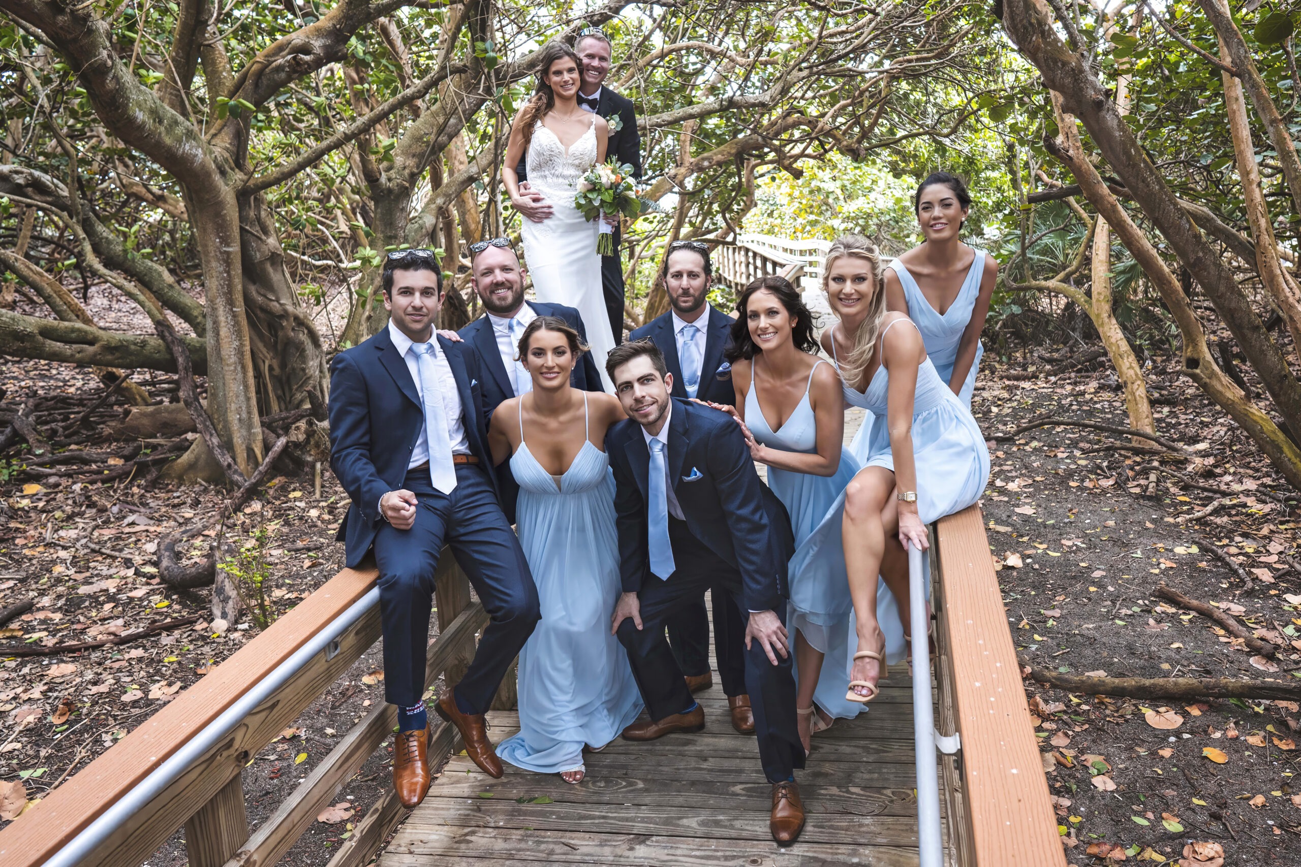 Bride and groom surrounded by their wedding party in light blue dresses and navy suits, posed along the wooden boardwalk under the lush sea grape canopy at Coral Cove Park on Jupiter Island, Florida. Captured by David Scarola Photography, this fine art wedding portrait blends natural beauty, joyful energy, and timeless elegance in one vibrant frame.