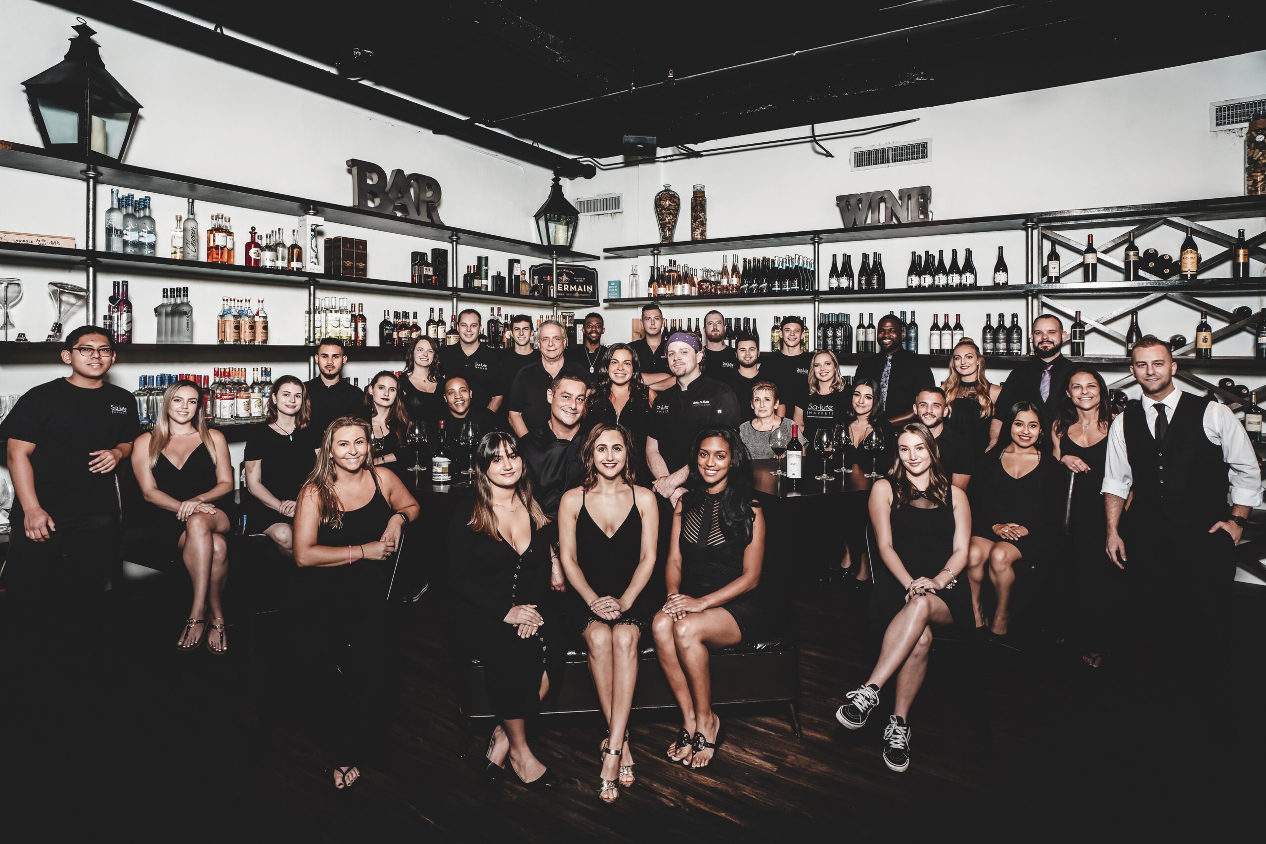 Full team portrait of Salute Market in Palm Beach Gardens, featuring restaurant, bar, and kitchen staff gathered together for a professional group photo inside their wine and spirits room.Salute Market Palm Beach Gardens, restaurant photography, bar staff photography, hospitality team portraits, kitchen staff photography, restaurant marketing photography, hospitality marketing, restaurant brand imagery, Palm Beach commercial photographer, South Florida restaurant content, David Scarola Photography, corporate photography Palm Beach, professional group photo, wine bar marketing, luxury restaurant portraits.