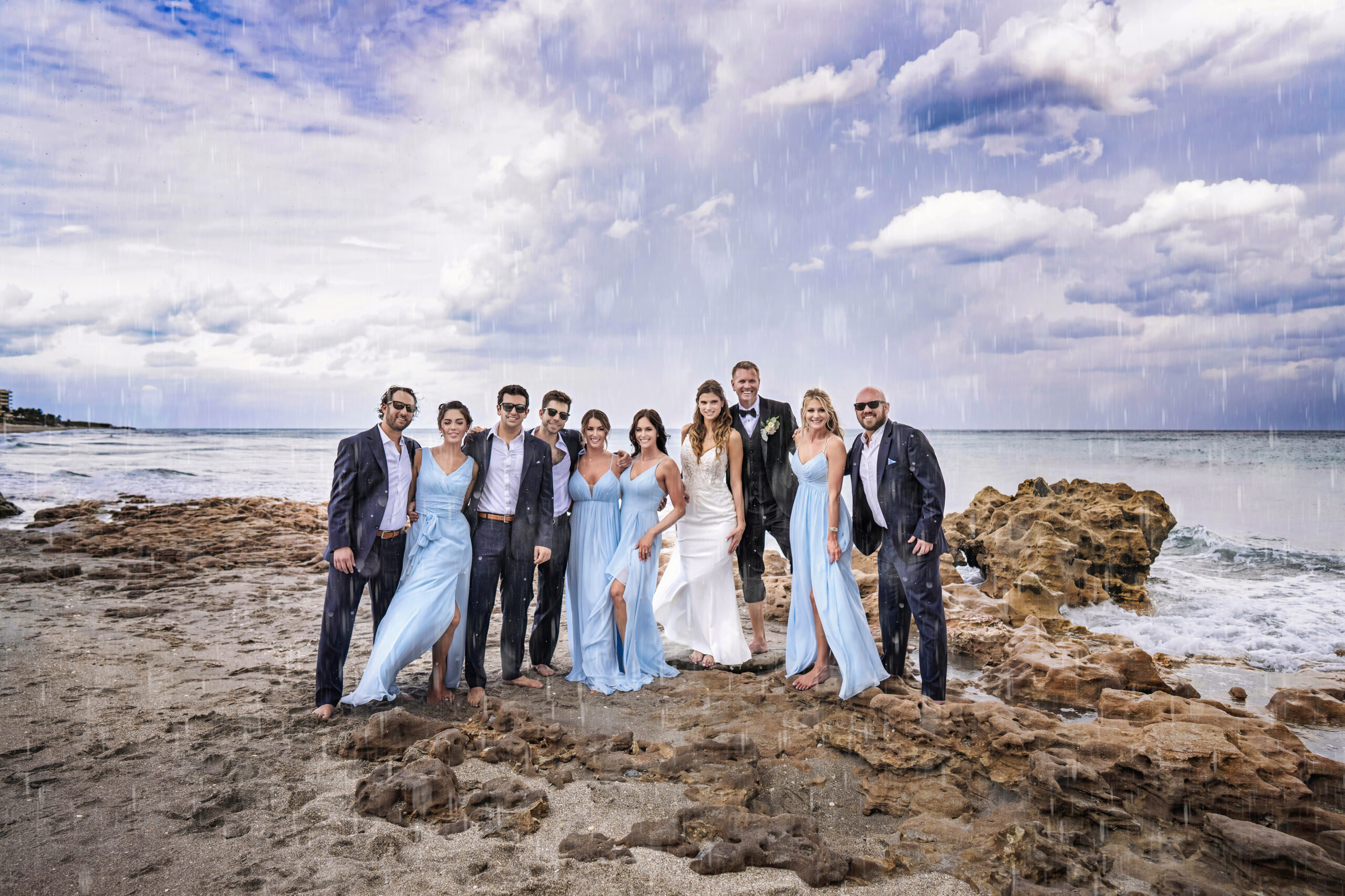 Wedding party on Jupiter Island standing barefoot on the rocky beach under dramatic clouds and light rain, dressed in elegant blue and white attire, capturing joy and beauty against the ocean horizon, having an absolute blast working with the best wedding photographer in the United States, David Scarola