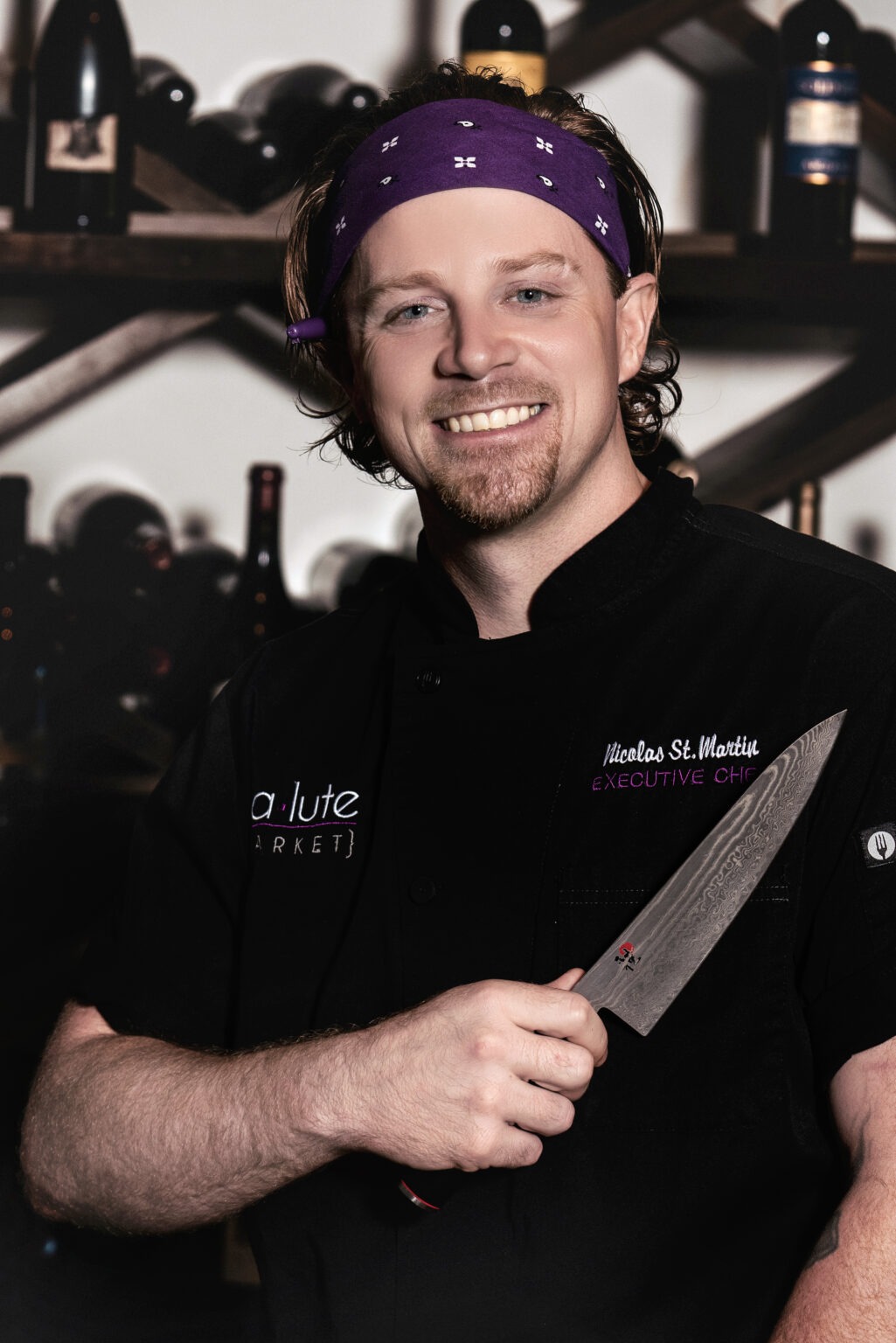 Executive Chef Nicolas St. Martin of Salute Market in Palm Beach Gardens, Florida, holding a chef’s knife and smiling confidently in the restaurant’s wine room. Professional chef portrait by David Scarola Photography.Executive Chef Nicolas St. Martin, Salute Market Palm Beach Gardens, restaurant photography Palm Beach, culinary portraits Florida, chef branding photography, professional chef portrait South Florida, hospitality photography Jupiter, restaurant marketing photography Palm Beach County, wine bar photography, David Scarola Photography, food and beverage photography, South Florida restaurant photography, Palm Beach Gardens executive chef, best restaurant photographer in Florida.