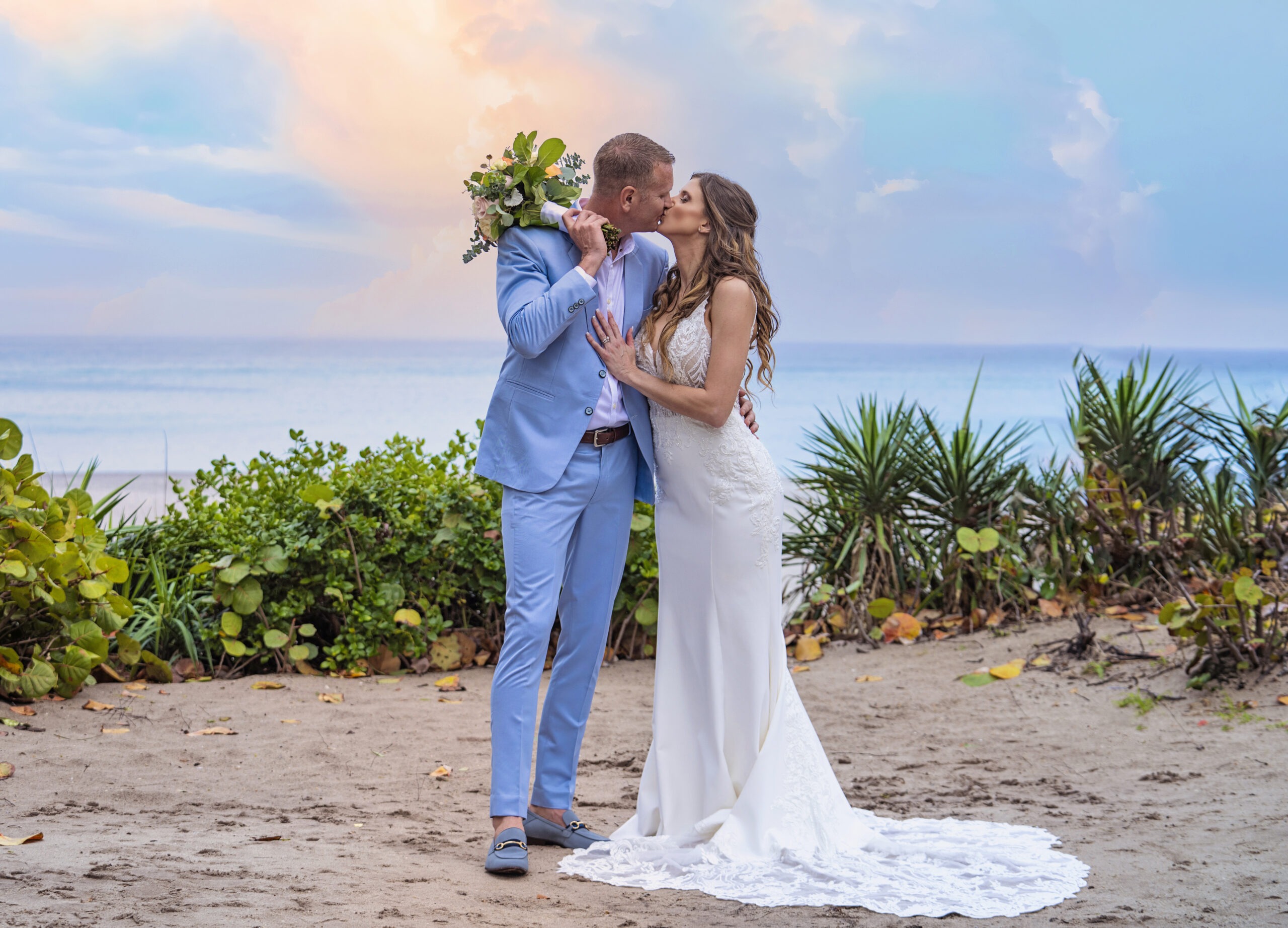 Bride and groom sharing a romantic kiss at their Jupiter Beach Resort wedding reception, framed by soft pastel skies and the tranquil Atlantic Ocean. The groom in a light blue suit and the bride in an elegant lace gown stand surrounded by tropical greenery — captured in luminous, fine-art detail by David Scarola, Florida’s best wedding photographer and master portrait artist specializing in coastal weddings across Palm Beach and Jupiter Island.