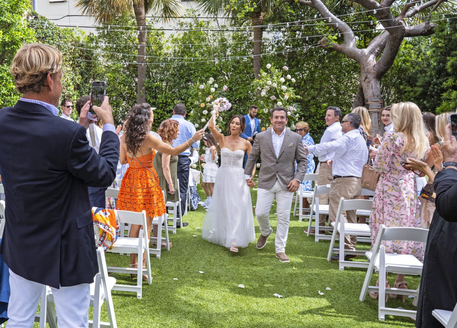 Just-married couple celebrating their walk down the aisle at The Colony Hotel, Palm Beach Island — surrounded by friends, florals, and tropical elegance. The bride raises her bouquet triumphantly as guests cheer under string lights. Captured by David Scarola, a Palm Beach wedding photographer celebrated for transforming real emotion into radiant, fine-art imagery filled with joy, color, and coastal sophistication.