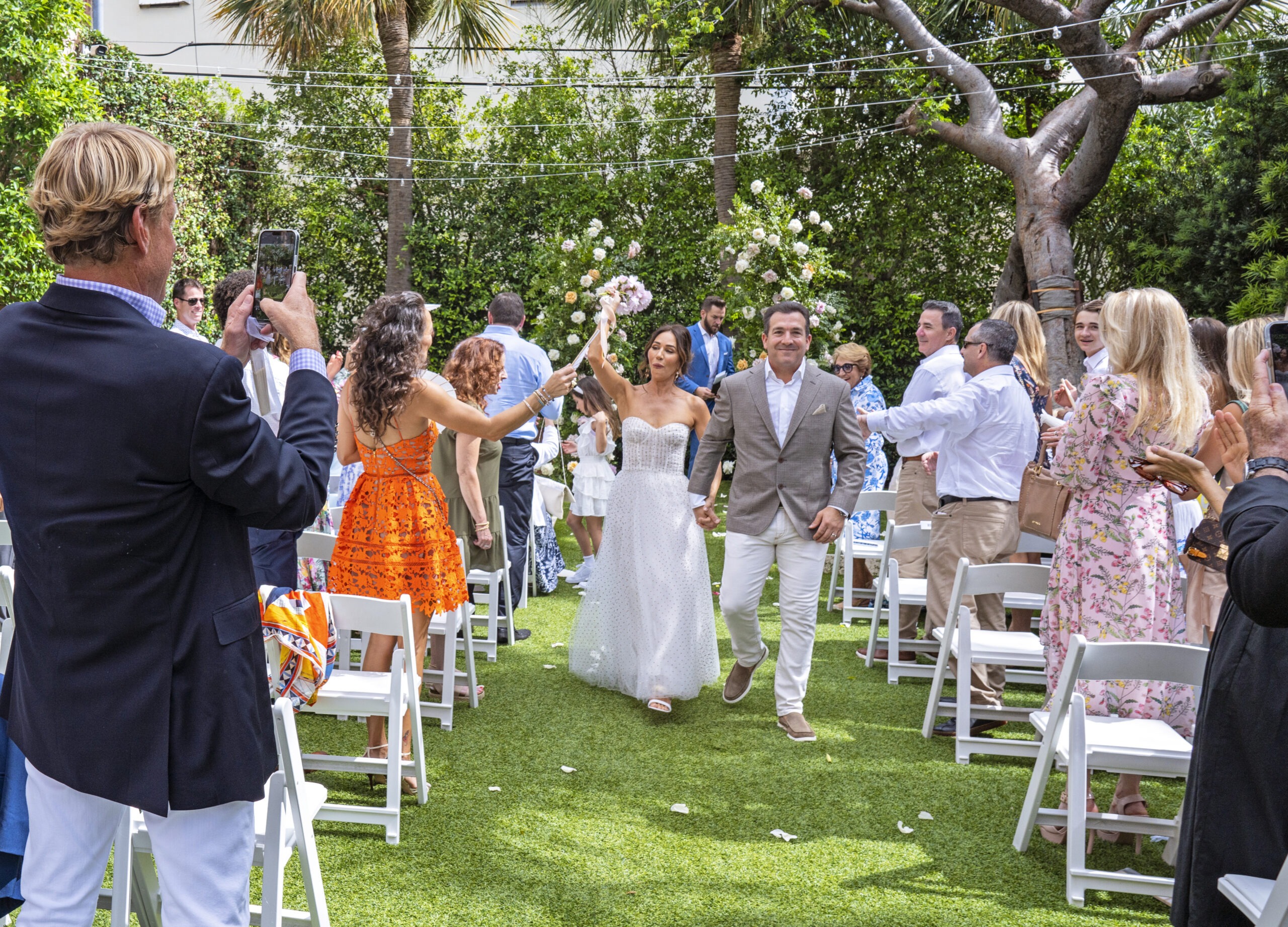 Just-married couple celebrating their walk down the aisle at The Colony Hotel, Palm Beach Island — surrounded by friends, florals, and tropical elegance. The bride raises her bouquet triumphantly as guests cheer under string lights. Captured by David Scarola, a Palm Beach wedding photographer celebrated for transforming real emotion into radiant, fine-art imagery filled with joy, color, and coastal sophistication.