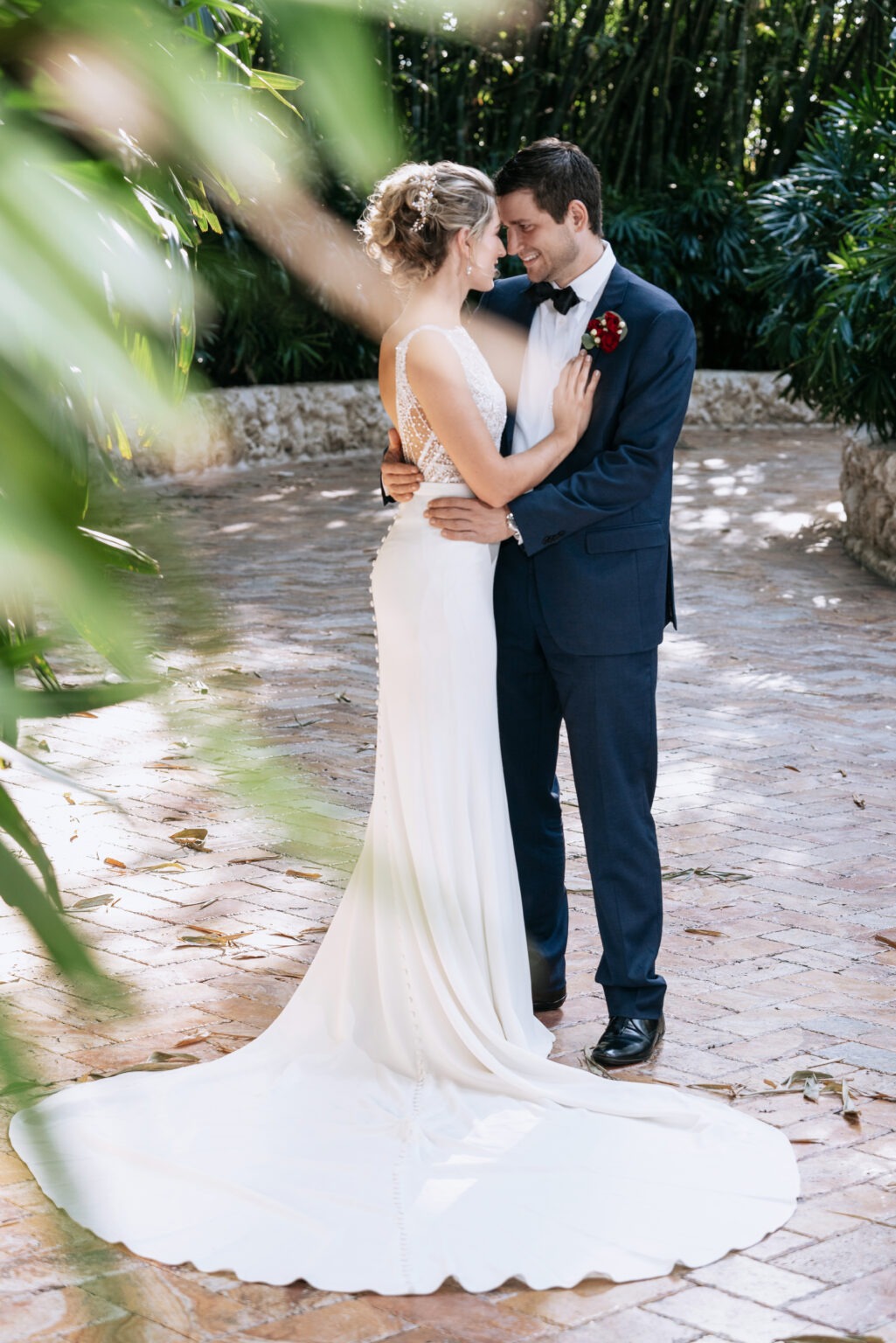 Bride and groom sharing an intimate moment surrounded by lush tropical greenery on Jupiter Island, Florida — her elegant gown flowing across the sunlit brick path as they embrace in quiet joy. Captured with depth, emotion, and artistry by David Scarola, Jupiter Island’s best wedding photographer and internationally recognized fine-art portrait artist known for natural light, timeless composition, and heartfelt storytelling.