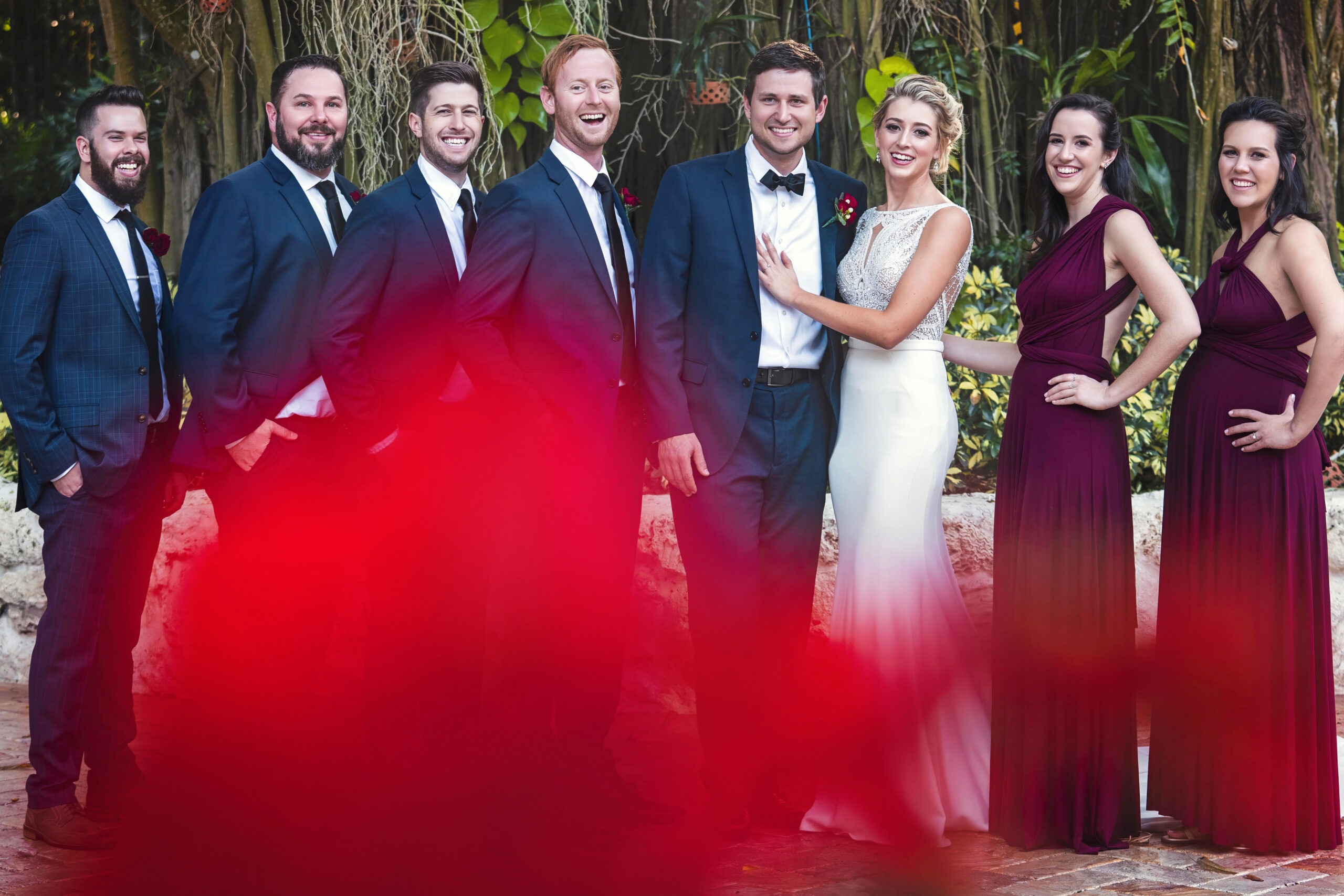 Elegant Jupiter Island wedding party portrait captured by Palm Beach wedding photographer David Scarola — bride and groom surrounded by bridesmaids in deep burgundy gowns and groomsmen in navy suits, framed by lush tropical greenery and a soft red floral foreground for a creative, artful touch.