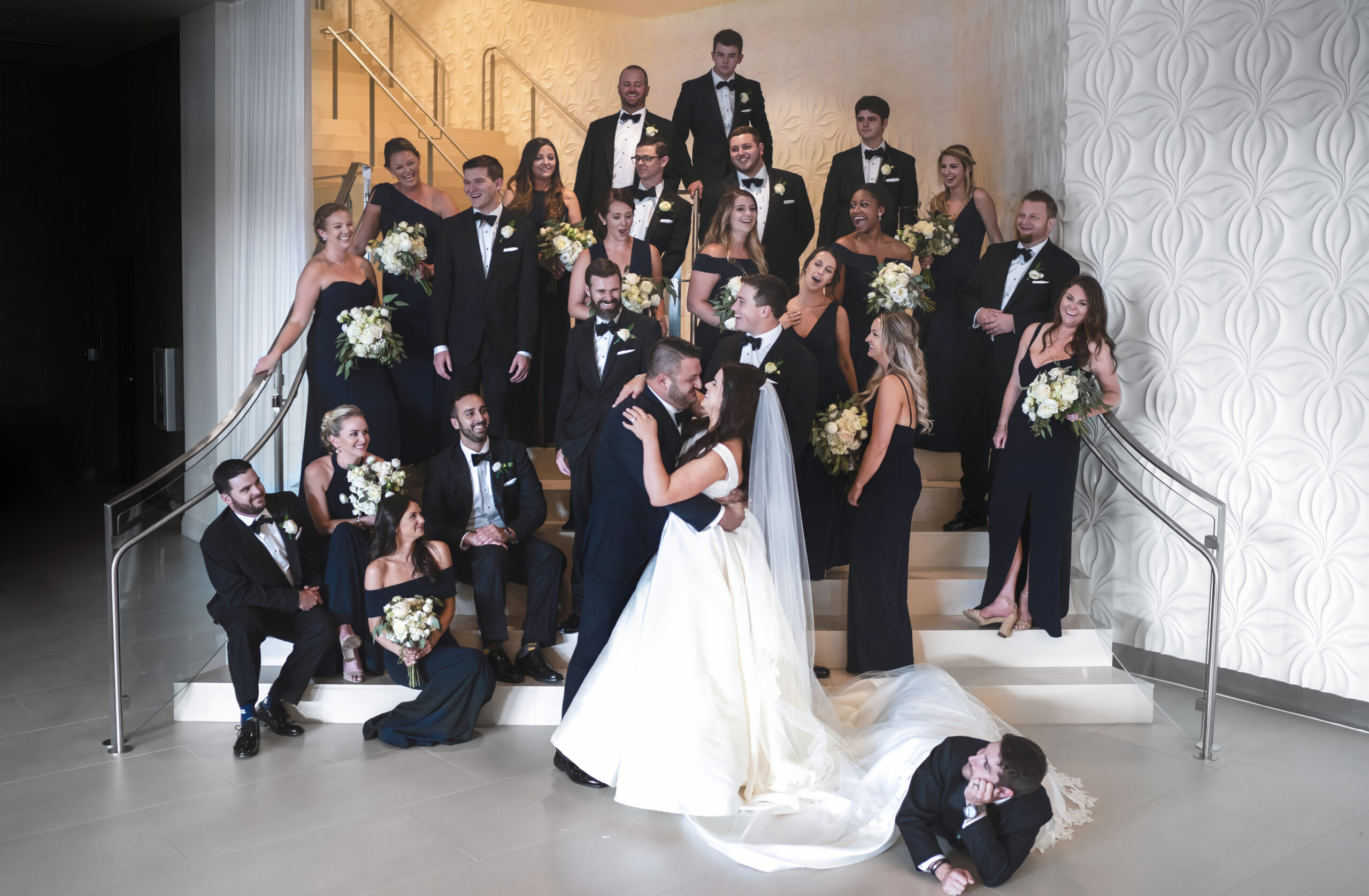 Large wedding party gathered on the elegant staircase of the Wyndham Grand at Harbourside, Jupiter Island — bridesmaids in navy, groomsmen in black tuxedos, and the couple sharing a romantic kiss at center — captured by David Scarola, Florida’s best wedding photographer and master fine-art portrait artist specializing in luxury weddings across Palm Beach and Jupiter.
