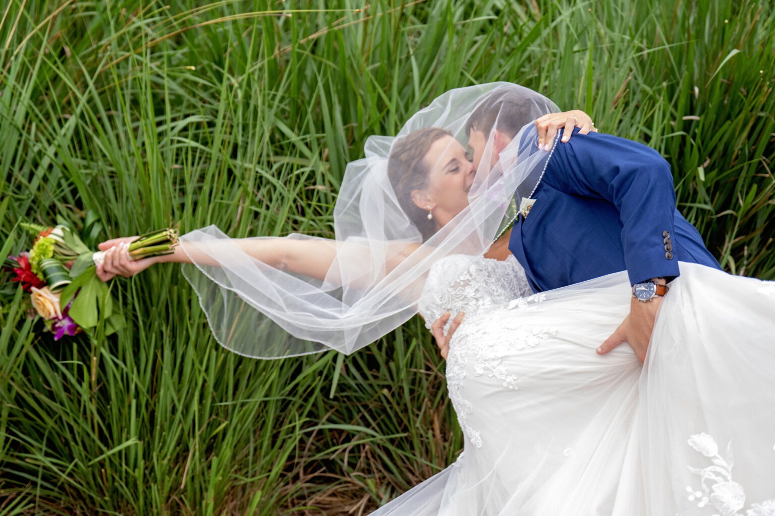 Bride and groom sharing a passionate kiss beneath her veil as he dips her gracefully, bouquet extended, surrounded by tall green reeds at Jupiter Country Club on Jupiter Island, Florida — captured in radiant fine-art detail by David Scarola, Florida’s best wedding photographer and internationally acclaimed portrait artist known for cinematic, emotional, and timeless wedding imagery.