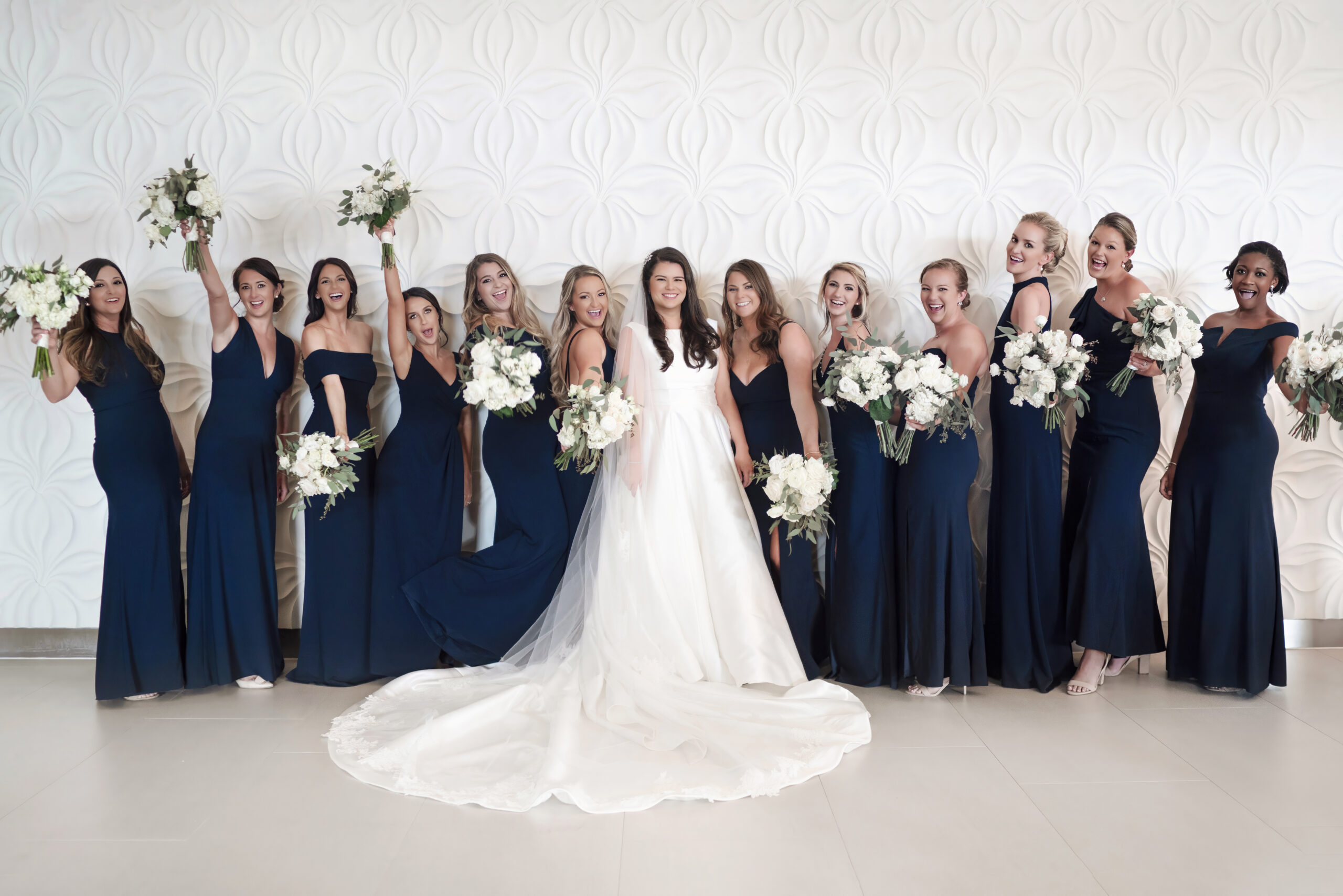Bride surrounded by her bridesmaids dressed in elegant navy gowns, all holding white floral bouquets and celebrating with joy inside the Wyndham Grand on Jupiter Island, Florida. Captured in bright, sophisticated detail by David Scarola, Florida’s best wedding photographer and master fine-art portrait artist known for luxurious, modern, and joy-filled wedding imagery.