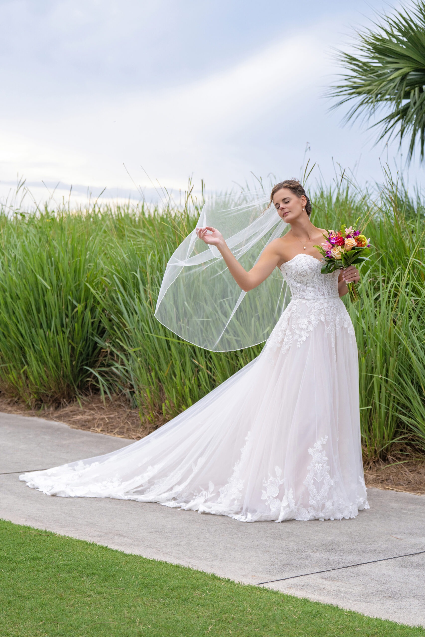 Stunning bride at Jupiter Country Club on Jupiter Island, Florida — standing gracefully in her lace gown with flowing veil and vibrant bouquet, surrounded by lush green reeds and soft coastal light. Captured in elegant fine-art style by David Scarola, Florida’s best wedding photographer and internationally acclaimed portrait artist known for luminous, romantic, and timeless wedding imagery.