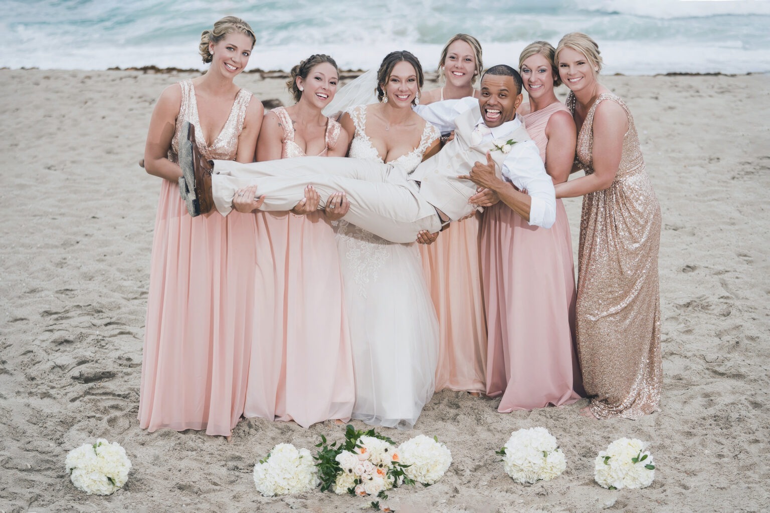 Bride and bridesmaids in blush and rose-gold dresses joyfully lifting the groom on the sandy shore of Palm Beach Island, Florida — ocean waves behind them, laughter and celebration in the air. Captured in vibrant fine-art detail by David Scarola, Florida’s best wedding photographer and master portrait artist known for timeless, joyful, and light-filled beach weddings.