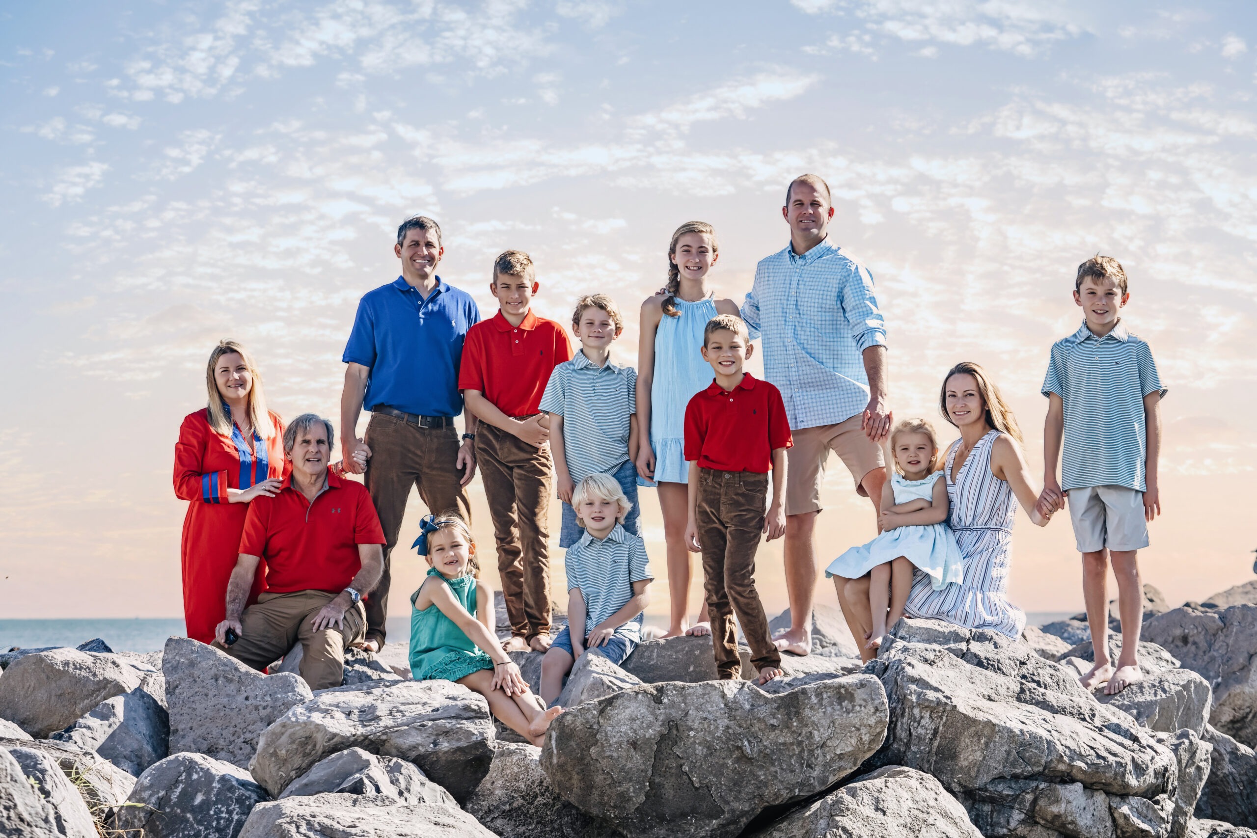An extended family of thirteen poses barefoot on coastal rocks beneath the soft morning sky at Bill Baggs Cape Florida Lighthouse in Key Biscayne. Dressed in coordinated hues of red, blue, and seafoam, they smile naturally toward the camera. Captured by David Scarola Photography, one of Florida’s premier family portrait artists known for blending fine art composition with coastal authenticity.David Scarola Photography, Key Biscayne family portraits, Bill Baggs Lighthouse photo shoot, extended family photography Florida, South Florida portrait photographer, Miami family photographer, Jupiter Island photographer, Palm Beach family photography, fine art family portraits, professional family photographer Florida, coastal family photo shoot, Florida beach portraits, master family photographer, top family photographers Palm Beach, Scarola Style photography, luxury portrait sessions Florida, family photography Key Biscayne lighthouse, natural light portraiture Florida, David Scarola family photographer, Jupiter Florida portraits.