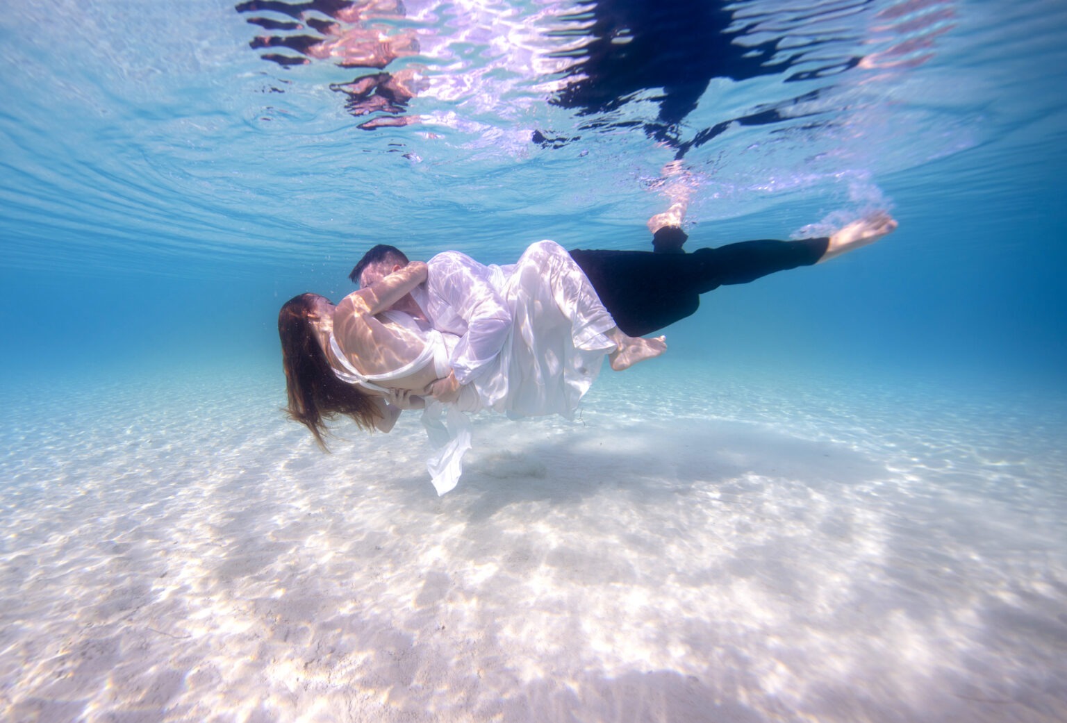 Underwater wedding portrait of a bride and groom embracing in crystal-clear turquoise water off Eleuthera, Bahamas. The couple floats weightlessly, wrapped in light and love, captured by David Scarola Photography, a fine-art destination wedding photographer specializing in underwater and creative beach weddings. This ethereal image blends romance, movement, and cinematic storytelling beneath the Bahamian sun.