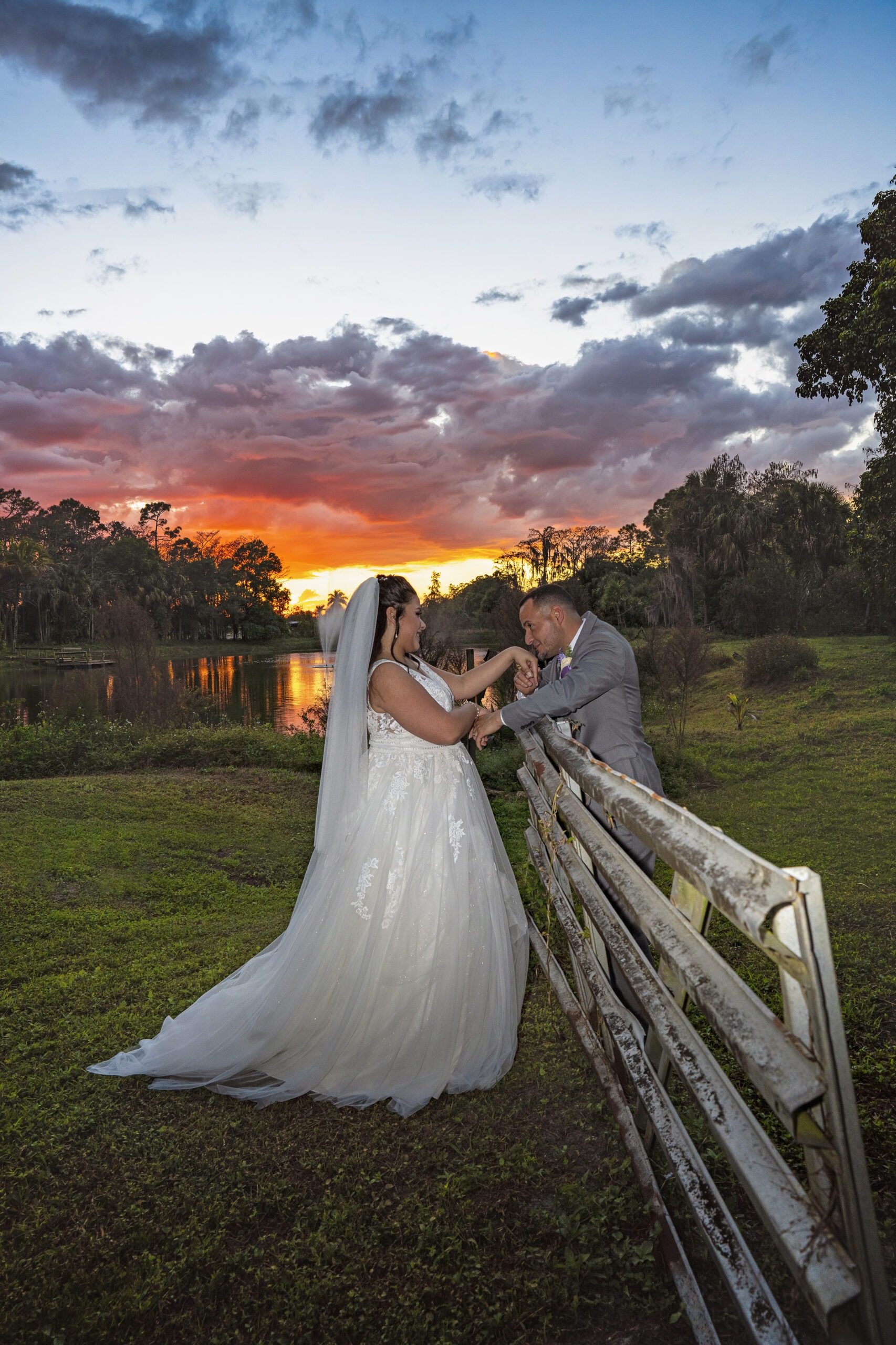 Bride and groom sharing a quiet, intimate moment by a rustic fence at Hawk’s Landing in Jupiter Farms, Florida — the sky ablaze with a magical sunset reflected over the lake. Captured in stunning natural light by David Scarola, Florida’s best wedding photographer and master fine-art portrait artist known for transforming fleeting moments into timeless works of emotion and beauty.