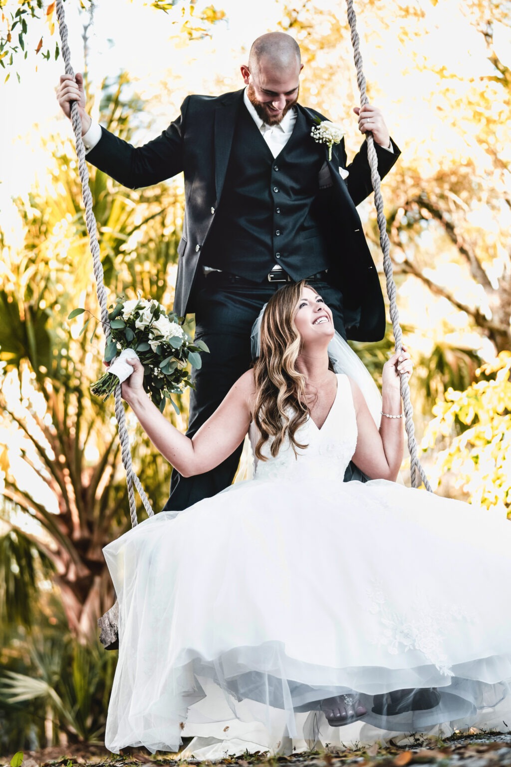Bride and groom sharing a joyful, intimate moment on a rustic swing surrounded by golden light and tropical palms on Jupiter Island, Florida — captured in radiant fine-art style by David Scarola, Florida’s best wedding photographer and master portrait artist known for his cinematic, romantic, and timeless wedding imagery.