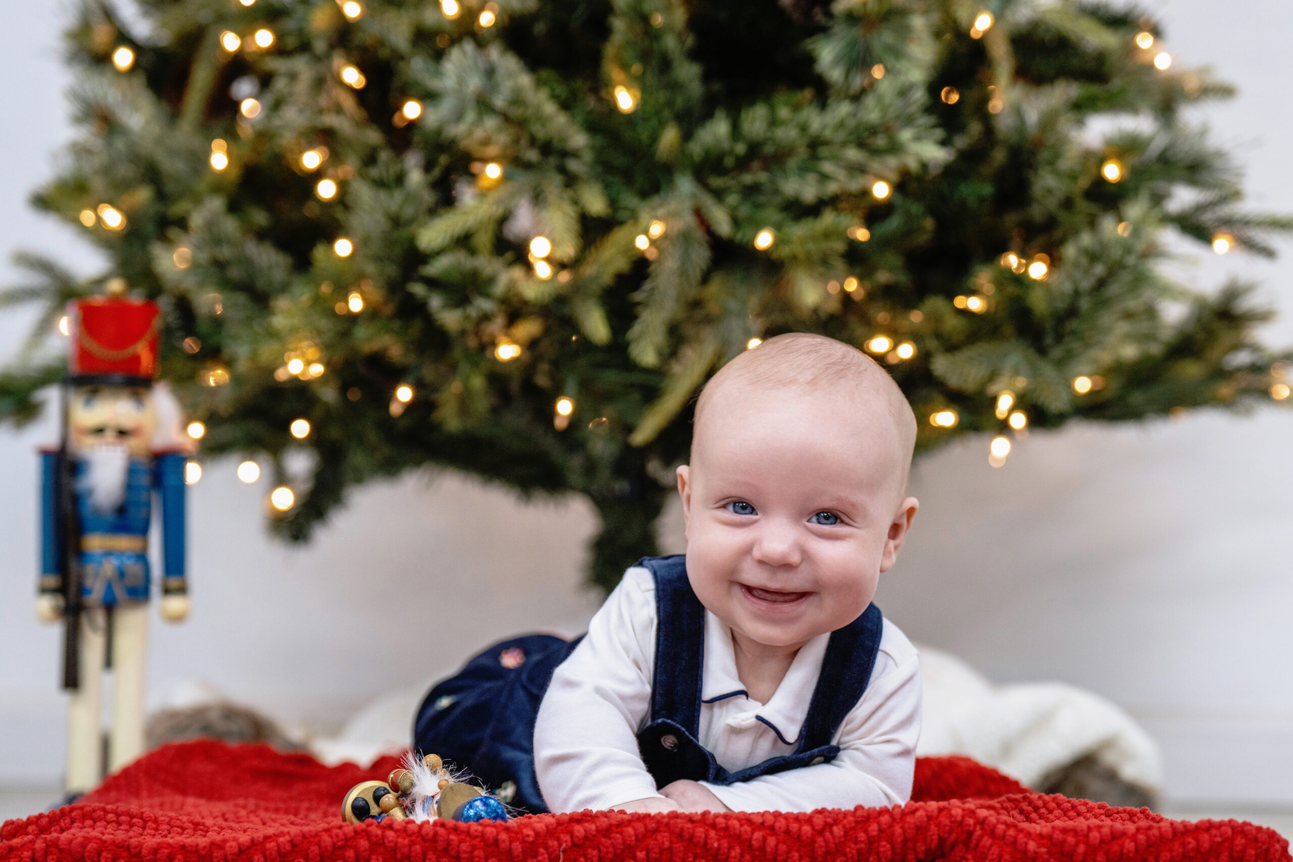 Smiling baby celebrating first Christmas in Palm Beach Island, lying on a red blanket beneath a decorated tree with twinkling lights and a nutcracker toy. Captured by David Scarola Photography, the image radiates joy, innocence, and holiday warmth — perfect Palm Beach family holiday portrait.